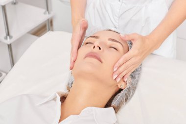 Güzellik uzmanı, güzellik salonunda gençleştirme için nemlendirici krem sürdükten sonra cildine masaj yapıyor, yaşlanma önleyici kozmetik prosedürler uyguluyor. Kozmetik uzmanı rahatlamak için kadın yüzüne masaj yapıyor.