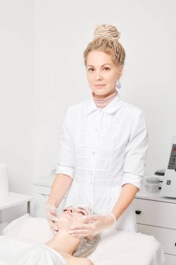 Kozmetik uzmanı, güzellik salonundaki yaşlanma karşıtı kozmetik prosedürleri ve cilt bakımı için kadın yüzüne krem masajı yapıyor. Eldivenli güzellik uzmanı elleri kadın yüzüne krem sürüyor.