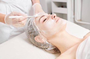 Güzellik uzmanı, güzellik salonlarında cilt bakımı için kadın yüzüne vitamin kremi maskesi, yaşlanma karşıtı kozmetik işlemler uyguluyor. Eldivenli kozmetik uzmanı maske takmak için kozmetik fırça tutuyor