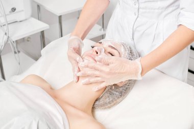 Kozmetik uzmanı, güzellik salonundaki yaşlanma karşıtı kozmetik prosedürleri ve cilt bakımı için kadın yüzüne krem masajı yapıyor. Eldivenli güzellik uzmanı elleri kadın yüzüne krem sürüyor.