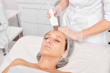 Güzellik uzmanı, güzellik salonundaki yaşlanma karşıtı kozmetik işlemleri ve cilt bakımını yenilemek için yüz ultrasoniği yapıyor. Kozmetik uzmanı yüz bakımını ultrason temizleyicisiyle yapıyor.