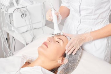 Güzellik uzmanı, güzellik salonundaki yaşlanma önleyici kozmetik işlemleri ve fonoforezi kullanarak gençleşen kadın yüzü için ultrason cildi sıkılaştırıyor. Kozmetik uzmanı ultrasonik deri kaldırıyor.