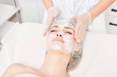 Kozmetik uzmanı, güzellik salonundaki yaşlanma karşıtı kozmetik prosedürleri ve cilt bakımı için kadın yüzüne krem masajı yapıyor. Eldivenli güzellik uzmanı elleri kadın yüzüne krem sürüyor.