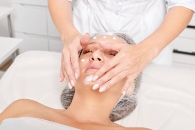 Güzellik salonunda kozmetik uzmanı cildine nemlendirici krem maskeyi sürüp gençleştirme, yaşlanmayı önleyici kozmetik prosedür uyguluyor. Güzellik uzmanı elleri kadın derisi için yüz kremi sürüyor.
