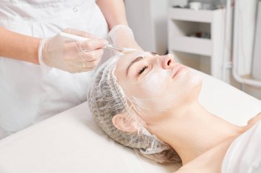 Güzellik uzmanı, güzellik salonundaki yaşlanma karşıtı kozmetik işlemler için cildini yenilemek için kadın yüzüne kozmetik krem maskesi sürüyor. Eldivenleri teslim eden kozmetik uzmanı maske takmak için kozmetik fırça tutuyor.