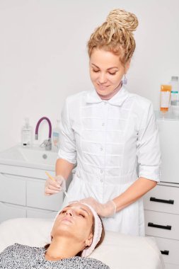 Kozmetik uzmanı, güzellik salonundaki kozmetik bakım ürünlerini nemlendirmek için kadın yüzüne estetik krem takıyor. Eldivenli güzellik uzmanı fırçayla kozmetik krem maskesi takıyor.