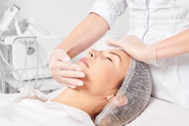 Güzellik uzmanı, güzellik salonunda gençleştirme için nemlendirici krem sürdükten sonra cildine masaj yapıyor, yaşlanma önleyici kozmetik prosedürler uyguluyor. Kozmetik uzmanı eldivenleri verdi. Rahatlamak için kadın yüzüne masaj yaptı.