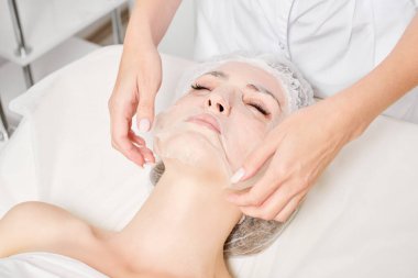 Güzellik uzmanı, güzellik salonundaki cilt bakımı için kadın yüzüne çarşaf maskesi, kırışıklık önleyici kozmetik prosedürler takıyor. Kozmetik uzmanı kadın yüzüne nemlendirici çarşaf maskesi uyguluyor.