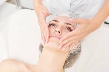 Güzellik uzmanları, güzellik salonlarında cilt bakımı, yaşlanma karşıtı estetik ameliyatlar için cilde masaj yapıyor. Kozmetik uzmanı güzel kadın yüzü için dairesel hareketlerle yüz masajı yapıyor.