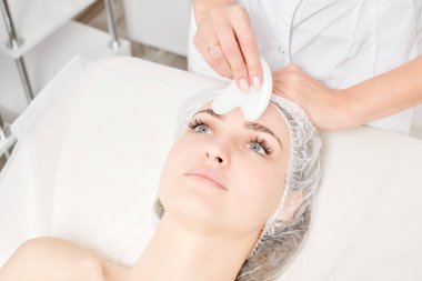 Gua Sha Taşı ile yüz masajı yapan kozmetik uzmanı, güzellik salonundaki lenfatik drenaj, yaşlanmayı önleyen estetik ameliyat için cilde bakıyor. Gua Sha aracıyla yüz masajı