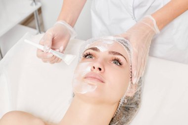 Güzellik uzmanı, güzellik salonlarında cilt bakımı için kadın yüzüne vitamin kremi maskesi, yaşlanma karşıtı kozmetik işlemler uyguluyor. Eldivenli kozmetik uzmanı maske takmak için kozmetik fırça tutuyor