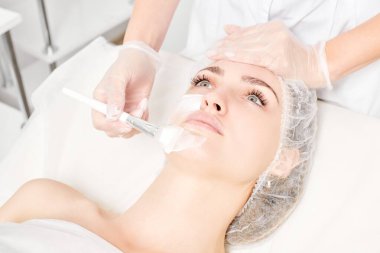 Güzellik uzmanı, güzellik salonlarında cilt bakımı için kadın yüzüne vitamin kremi maskesi, yaşlanma karşıtı kozmetik işlemler uyguluyor. Eldivenli kozmetik uzmanı maske takmak için kozmetik fırça tutuyor