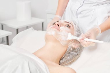Güzellik uzmanı, güzellik salonundaki yaşlanma karşıtı kozmetik işlemler için cildini yenilemek için kadın yüzüne kozmetik krem maskesi sürüyor. Eldivenleri teslim eden kozmetik uzmanı maske takmak için kozmetik fırça tutuyor.
