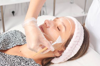 Güzellik uzmanı, güzellik salonundaki kozmetik krem maskesini cilt tazeleme, nemlendirici kozmetik işlemler için kadın yüzüne uyguluyor. Eldivenli kozmetik uzmanı fırçayla kozmetik krem maskesi takıyor.