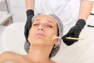 Güzellik uzmanı kadın yüzüne nemlendirici yüz derisi ve güzellik salonundaki yaşlanma karşıtı kozmetik prosedürleri için bal maskesi takıyor. Siyah eldivenli kozmetik uzmanı bal maskesi takmak için kozmetik fırça tutuyor.