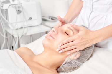 Güzellik salonunda kozmetik uzmanı cildine nemlendirici krem maskeyi sürüp gençleştirme, yaşlanmayı önleyici kozmetik prosedür uyguluyor. Güzellik uzmanı elleri kadın derisi için yüz kremi sürüyor.