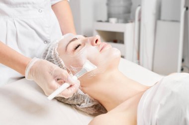 Kozmetik uzmanı kadın yüzüne cilt tazeleme için krem maskesi, güzellik salonundaki yaşlanma karşıtı kozmetik prosedürleri uyguluyor. Eldivenli güzellik uzmanı elleri kozmetik fırça tutuyor ve kozmetik maske takıyor