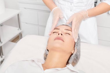 Güzellik uzmanı, güzellik salonunda gençleştirme için nemlendirici krem sürdükten sonra cildine masaj yapıyor, yaşlanma önleyici kozmetik prosedürler uyguluyor. Kozmetik uzmanı eldivenleri verdi. Rahatlamak için kadın yüzüne masaj yaptı.