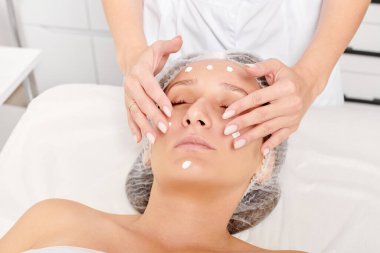 Güzellik uzmanları, güzellik salonlarında gençleştirme için nemlendirici krem maskeyi kadın yüzüne sürerler, yaşlanma karşıtı kozmetik işlemler yaparlar. Kozmetik uzmanı kadın derisi için yüz kremi uyguluyor.