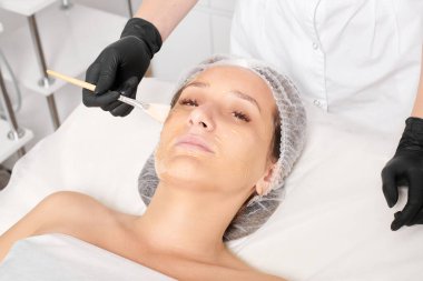 Kozmetik uzmanı kadın yüzüne nemlendirici yüz derisi ve güzellik salonundaki yaşlanma karşıtı kozmetik prosedürü için bal maskesi takıyor. Siyah eldivenli güzellik uzmanı, bal maskesi takmak için kozmetik fırça tutuyor.