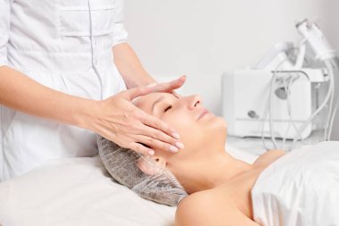 Güzellik salonunda kozmetik uzmanı cildine nemlendirici krem maskeyi sürüp gençleştirme, yaşlanmayı önleyici kozmetik prosedür uyguluyor. Güzellik uzmanı elleri kadın derisi için yüz kremi sürüyor.