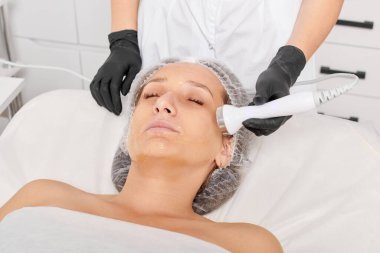 Güzellik uzmanı, güzellik salonundaki yaşlanma önleyici kozmetik işlemleri ve fonoforezi kullanarak gençleşen kadın yüzü için ultrason cildi sıkılaştırıyor. Kozmetik uzmanı ultrasonik deri kaldırıyor.