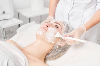 Güzellik uzmanı, güzellik salonundaki yaşlanma karşıtı kozmetik işlemler için cildini yenilemek için kadın yüzüne kozmetik krem maskesi sürüyor. Eldivenleri teslim eden kozmetik uzmanı maske takmak için kozmetik fırça tutuyor.