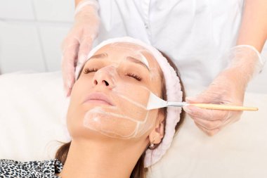 Kozmetik uzmanı, güzellik salonundaki kozmetik bakım ürünlerini nemlendirmek için kadın yüzüne estetik krem takıyor. Eldivenli güzellik uzmanı fırçayla kozmetik krem maskesi takıyor.