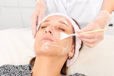 Güzellik uzmanı, güzellik salonundaki kozmetik krem maskesini cilt tazeleme, nemlendirici kozmetik işlemler için kadın yüzüne uyguluyor. Eldivenli kozmetik uzmanı fırçayla kozmetik krem maskesi takıyor.