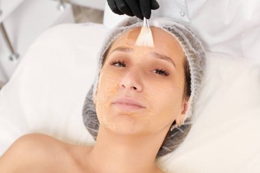 Güzellik uzmanı kadın yüzüne nemlendirici yüz derisi ve güzellik salonundaki yaşlanma karşıtı kozmetik prosedürleri için bal maskesi takıyor. Siyah eldivenli kozmetik uzmanı bal maskesi takmak için kozmetik fırça tutuyor.