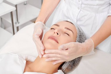 Kozmetik uzmanı, güzellik salonundaki gençleştirme için nemlendirici krem sürdükten sonra cildine masaj yapıyor. Eldivenli güzellik uzmanı elleri rahatlamak için kadın yüzüne masaj yapıyor.