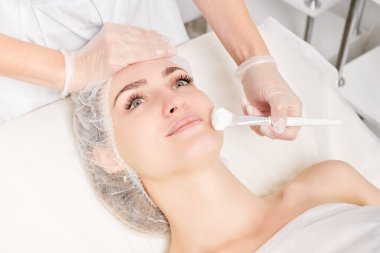 Güzellik uzmanı, güzellik salonundaki gençleşme yüzü için kadın yüzüne krem maske takar, yaşlanma karşıtı kozmetik işlemler yapar. Eldivenli kozmetik uzmanı kozmetik fırçasını tutuyor ve canlandırıcı maske takıyor.