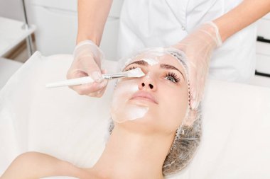 Güzellik uzmanı, güzellik salonundaki yaşlanma karşıtı kozmetik işlemler için cildini yenilemek için kadın yüzüne kozmetik krem maskesi sürüyor. Eldivenleri teslim eden kozmetik uzmanı maske takmak için kozmetik fırça tutuyor.