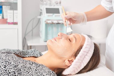 Güzellik uzmanı, güzellik salonundaki kozmetik krem maskesini cilt tazeleme, nemlendirici kozmetik işlemler için kadın yüzüne uyguluyor. Eldivenli kozmetik uzmanı fırçayla kozmetik krem maskesi takıyor.