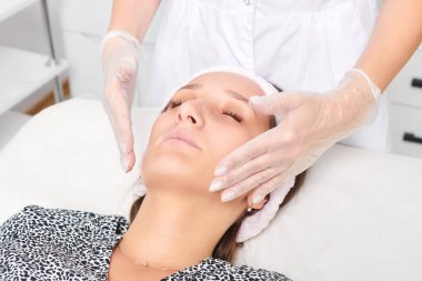 Eldivenli kozmetik uzmanı yenilenme, lenfatik drenaj, güzellik salonundaki krem cilt estetiği için kadın yüz masajı yapıyor. Güzellik uzmanı elleriyle cildini düzleştiriyor.