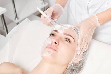 Güzellik uzmanı, güzellik salonundaki yaşlanma karşıtı kozmetik işlemler için cildini yenilemek için kadın yüzüne kozmetik krem maskesi sürüyor. Eldivenleri teslim eden kozmetik uzmanı maske takmak için kozmetik fırça tutuyor.