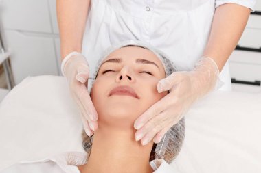 Güzellik uzmanı, güzellik salonunda gençleştirme için nemlendirici krem sürdükten sonra cildine masaj yapıyor, yaşlanma önleyici kozmetik prosedürler uyguluyor. Kozmetik uzmanı eldivenleri verdi. Rahatlamak için kadın yüzüne masaj yaptı.