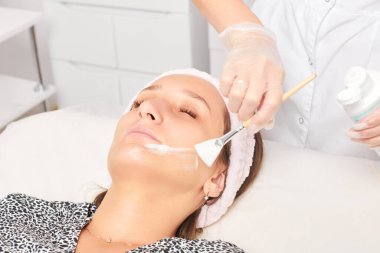 Kozmetik uzmanı, güzellik salonundaki kozmetik bakım ürünlerini nemlendirmek için kadın yüzüne estetik krem takıyor. Eldivenli güzellik uzmanı fırçayla kozmetik krem maskesi takıyor.