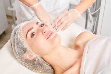 Kozmetik uzmanı kadın yüzüne gençleştirme enjeksiyonu yapıyor. Güzellik salonunda yaşlanmayı önleyen estetik cerrahi olmayan prosedür uyguluyor. Eldiven uzmanları şırınga ile iğne yapıyor.