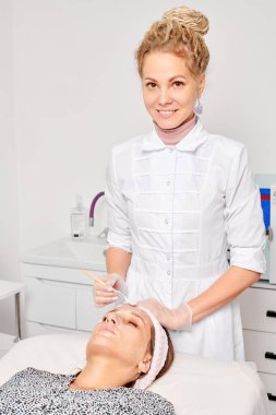 Güzellik uzmanları, güzellik salonundaki estetik ameliyatları nemlendiren cilt yenileme için kadın yüzüne kozmetik krem maskesi uyguluyor. Eldivenli kozmetik uzmanı fırçayla kozmetik krem maskesi takıyor.