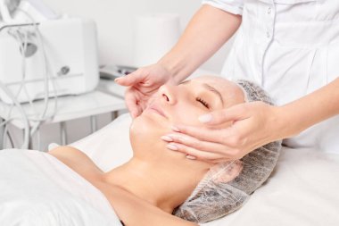 Güzellik salonunda kozmetik uzmanı cildine nemlendirici krem maskeyi sürüp gençleştirme, yaşlanmayı önleyici kozmetik prosedür uyguluyor. Güzellik uzmanı elleri kadın derisi için yüz kremi sürüyor.