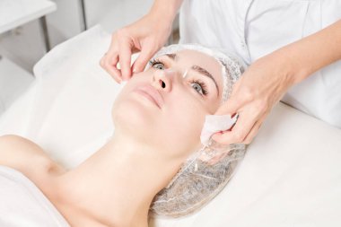 Güzellik uzmanı, güzellik salonundaki yüz kremi maskesini kadın yüzünden kozmetik mendillerle alıyor, cilt yenileme estetik ameliyatı yapıyor. Kozmetik uzmanı yüz maskesini çıkarmak için peçete tutuyor.