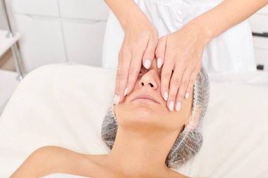 Kozmetik uzmanı, güzellik salonundaki cilt bakımı, yaşlanmayı önleyen estetik ameliyatlar için cilde masaj yapıyor. Güzellik uzmanı güzel kadın yüzü için dairesel hareketlerle yüz masajı yapıyor.