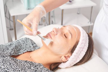 Kozmetik uzmanı, güzellik salonundaki kozmetik bakım ürünlerini nemlendirmek için kadın yüzüne estetik krem takıyor. Eldivenli güzellik uzmanı fırçayla kozmetik krem maskesi takıyor.