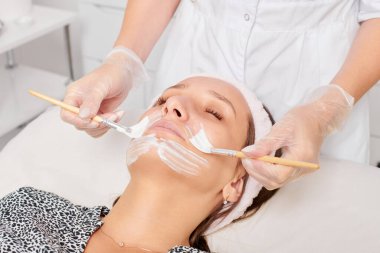Güzellik uzmanı, güzellik salonunda estetik krem maskesini cilt tazeleme, nemlendirici kozmetik işlemleri için kadın yüzüne uyguluyor. Eldivenli kozmetik uzmanı iki fırçayla kozmetik krem maskesi takıyor.