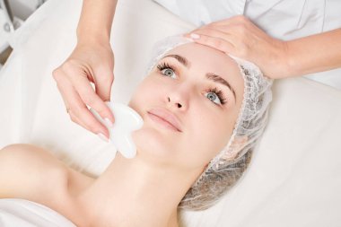 Gua Sha Taşı ile yüz masajı yapan kozmetik uzmanı, güzellik salonundaki lenfatik drenaj, yaşlanmayı önleyen estetik ameliyat için cilde bakıyor. Gua Sha aracıyla yüz masajı