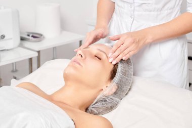 Güzellik salonunda kozmetik uzmanı cildine nemlendirici krem maskeyi sürüp gençleştirme, yaşlanmayı önleyici kozmetik prosedür uyguluyor. Güzellik uzmanı elleri kadın derisi için yüz kremi sürüyor.