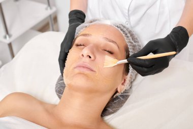 Kozmetik uzmanı kadın yüzüne nemlendirici yüz derisi ve güzellik salonundaki yaşlanma karşıtı kozmetik prosedürü için bal maskesi takıyor. Siyah eldivenli güzellik uzmanı, bal maskesi takmak için kozmetik fırça tutuyor.