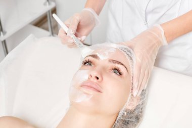 Güzellik uzmanı, güzellik salonlarında cilt bakımı için kadın yüzüne vitamin kremi maskesi, yaşlanma karşıtı kozmetik işlemler uyguluyor. Eldivenli kozmetik uzmanı maske takmak için kozmetik fırça tutuyor
