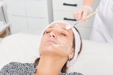 Güzellik uzmanı, güzellik salonundaki kozmetik krem maskesini cilt tazeleme, nemlendirici kozmetik işlemler için kadın yüzüne uyguluyor. Eldivenli kozmetik uzmanı fırçayla kozmetik krem maskesi takıyor.